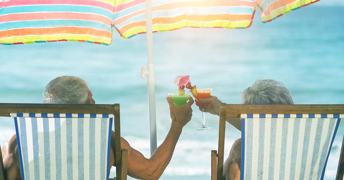 Seniorenpaar sitzt in Klappstühlen am Strand unter einem bunten Sonnenschirm und genießt Cocktails | Wohnen im Alter