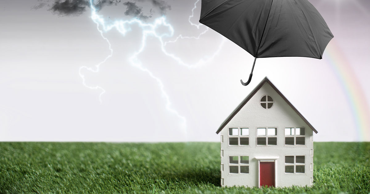 Haus auf einer grünen Wiese, dass vor Wetterergnissen und anderen Schadensfällen durch einen schwarzen Regenschirm geschützt wird.